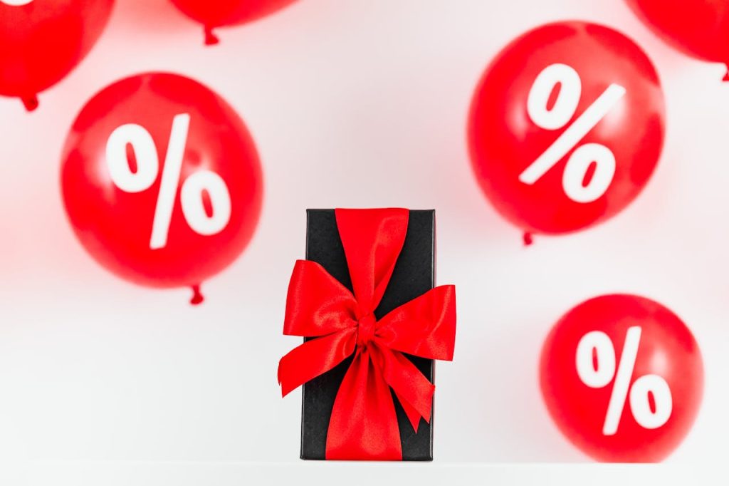 Bright red balloons with percentage signs surrounding a black gift box with a red ribbon on a white background.