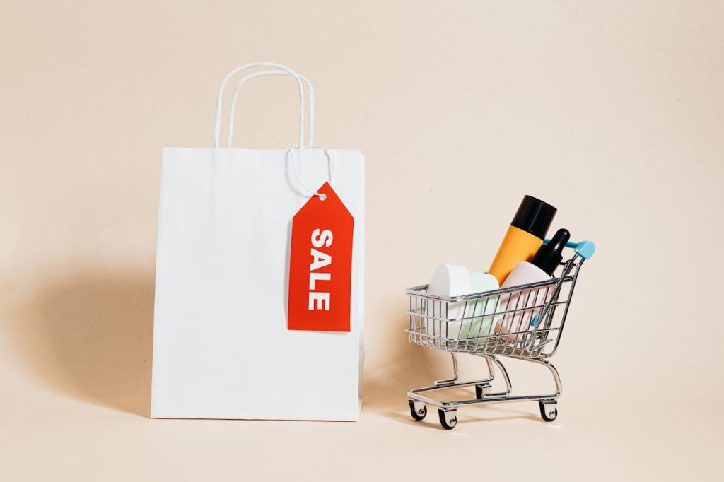 White shopping bag with red sale tag and mini shopping cart on beige background.