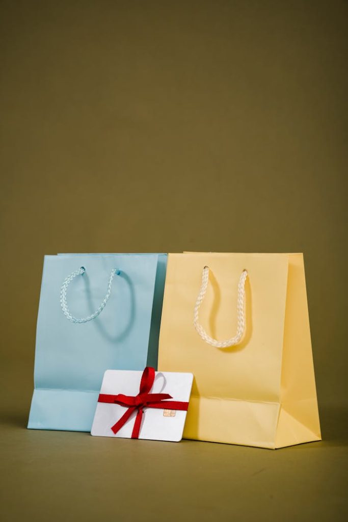 Pastel blue and yellow shopping bags with a red-ribboned gift card on an olive background, ideal for retail themes.